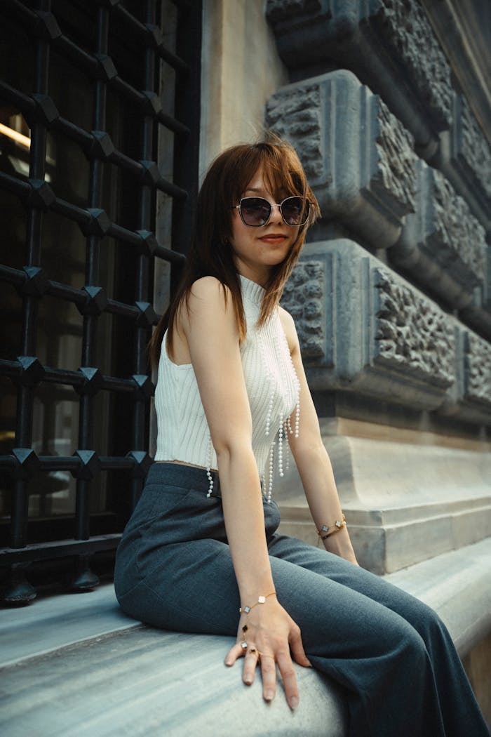 Elegant woman with sunglasses relaxing on a stone ledge outdoors. Urban fashion vibe.